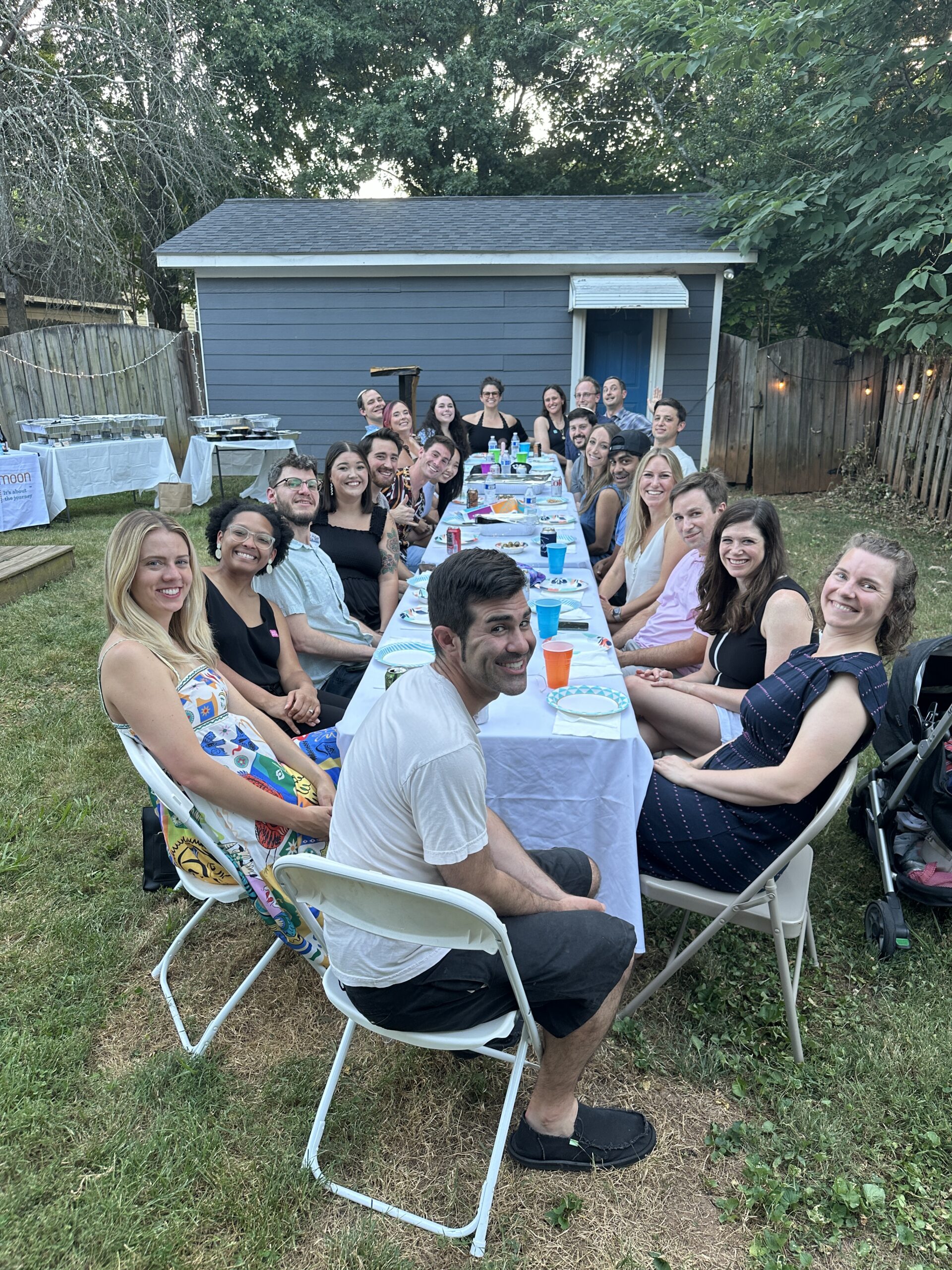 People are gathered outdoors, seated around a long table enjoying a meal together.