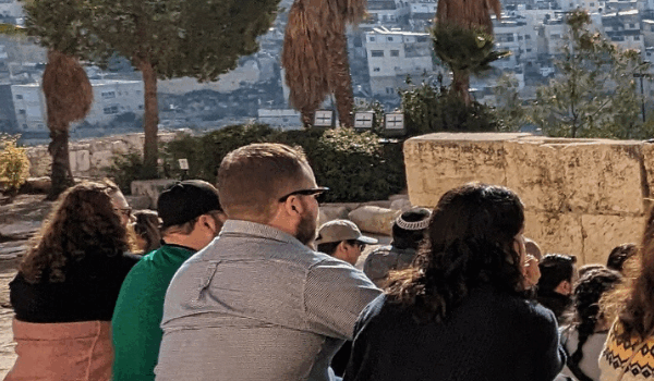 A group of people sitting outdoors near ancient stone ruins, possibly on a tour or visit, with a cityscape in the background.
