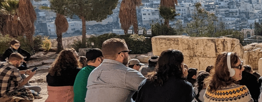A group of people sitting outdoors near ancient stone ruins, possibly on a tour or visit, with a cityscape in the background.