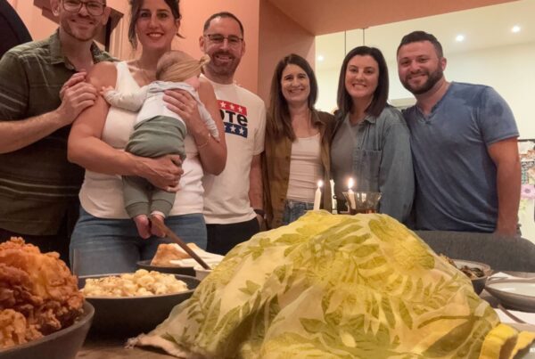 A group of smiling people poses with a baby around a table with food, candles, and a cloth-covered dish.