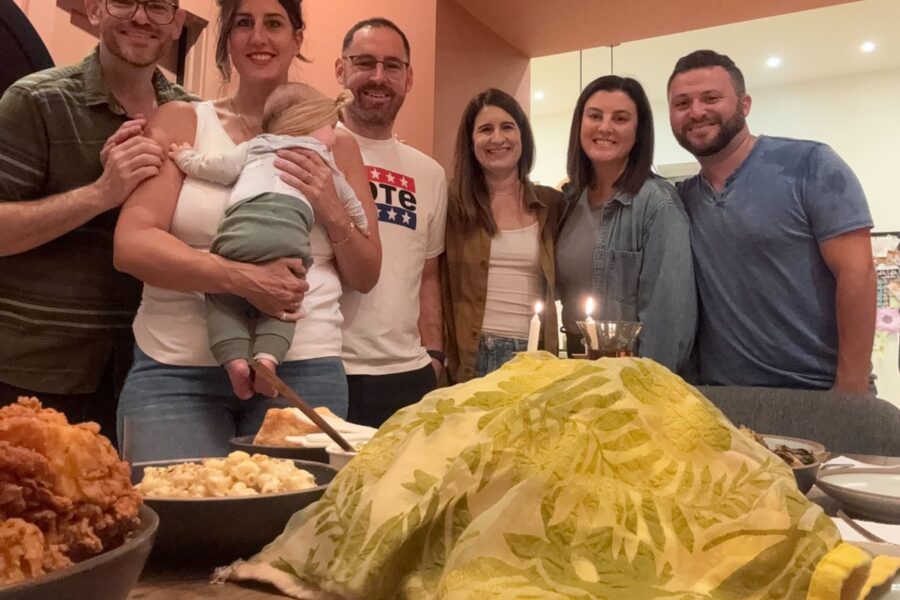 A group of smiling people poses with a baby around a table with food, candles, and a cloth-covered dish.