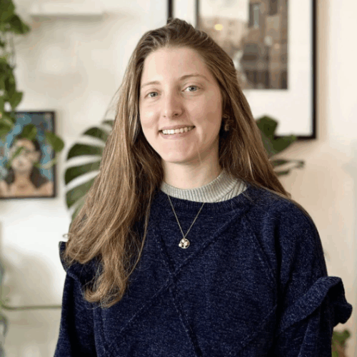 A smiling person with long hair and a blue sweater stands indoors, surrounded by artwork and plants.