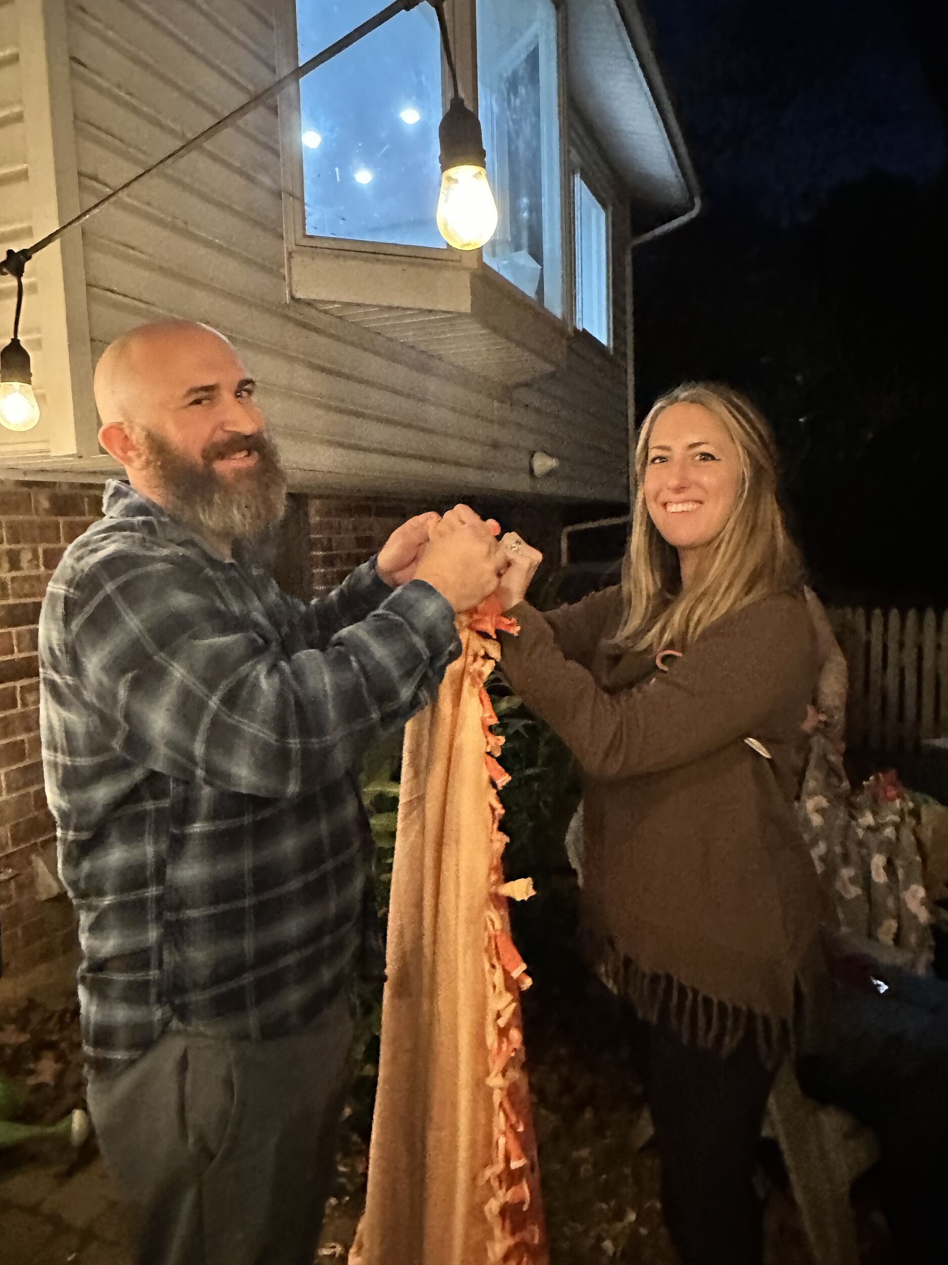A man and a woman are outdoors, smiling and holding a fringed blanket together. String lights illuminate the scene.